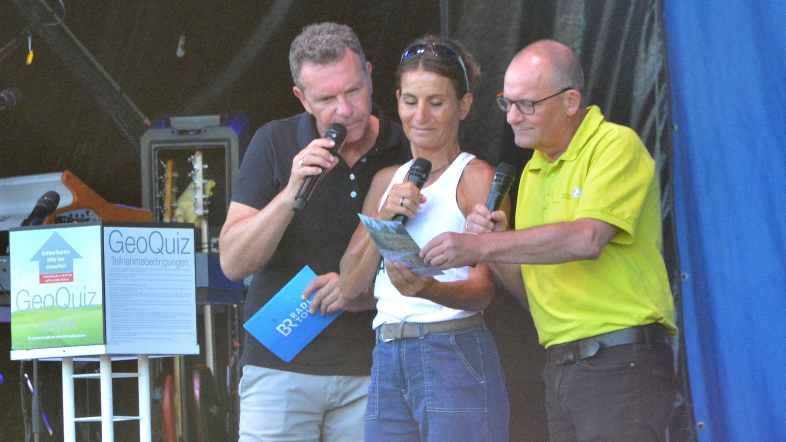 LDBV-Vizepräsident Oliver Schrempel (rechts) gemeinsam mit BR-Moderator Roman Roell und Karina Ladwig bei der Auslosung des Gewinners des täglichen Geoquiz Drei Menschen mit Mikrofonen
